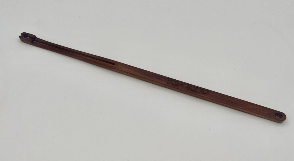 A long, slender wooden stick engraved with "Ipe," featuring a small notch and hole at one end, rests on a plain white surface.