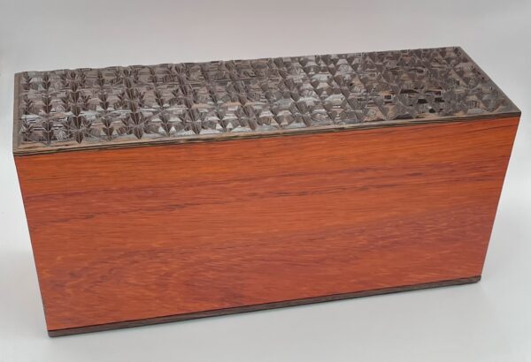 Hideaway is a rectangular wooden box with smooth reddish-brown sides and a dark, intricately carved lid featuring a raised geometric pattern. It is pictured against a plain light background.