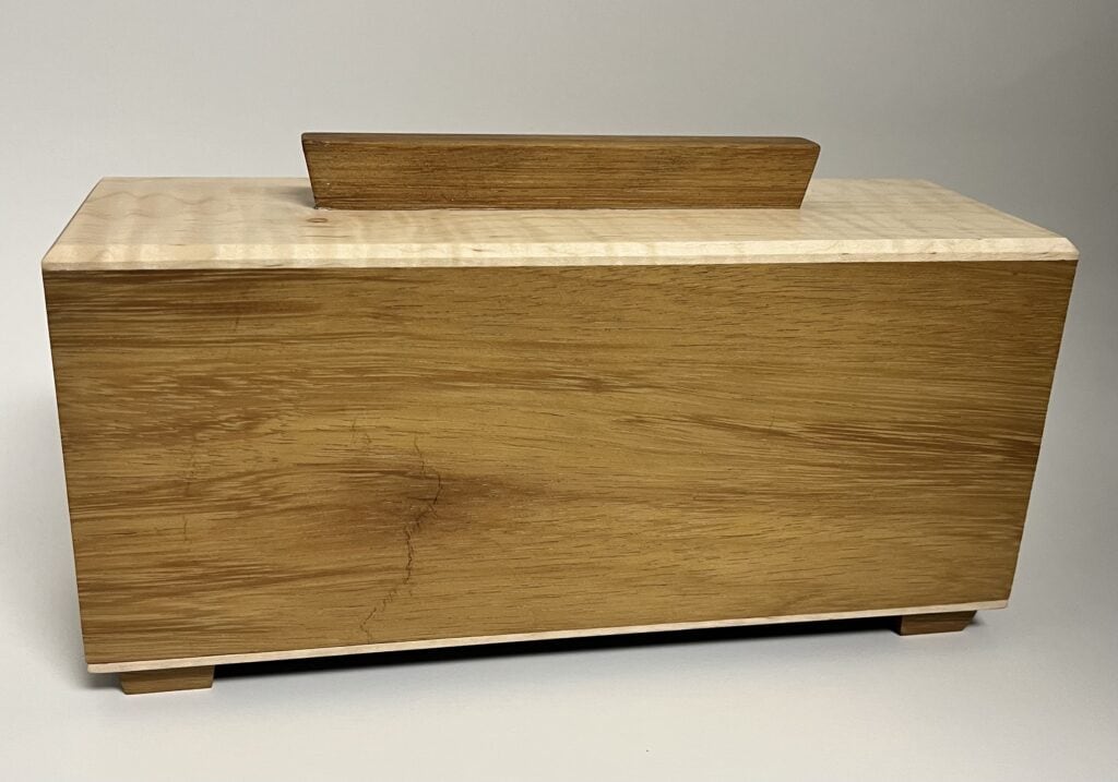 The Hideaway is a rectangular wooden box with a smooth, light brown finish, featuring a flat lid with a central handle and four small feet, set against a plain background.