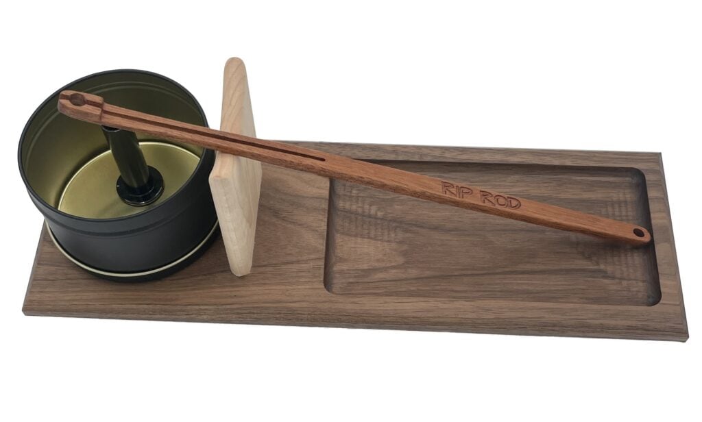 The Rip Rod Essentials features a wooden base holding a handled metal tin, a vertical wooden block, and a long engraved "RIP ROD" stick, all neatly arranged on the rectangular tray.