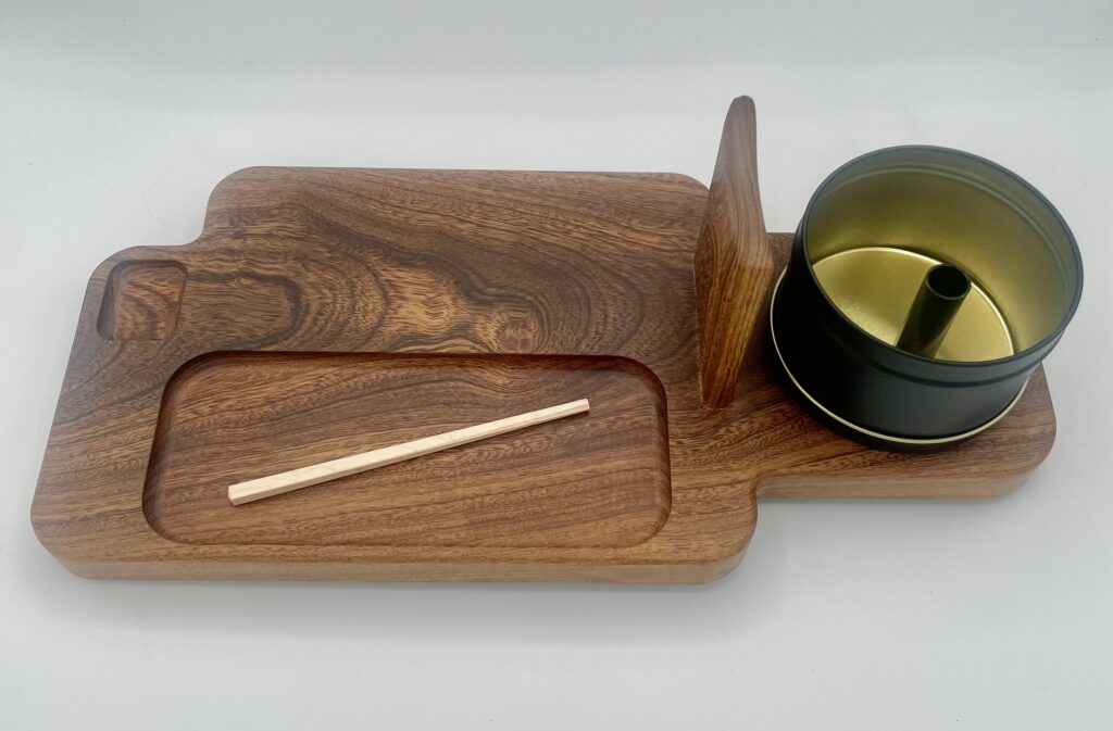 A wooden serving tray with a carved rectangular section, a groove for holding items, a single chopstick, and a round black and gold metal container with a center divider on the right side.